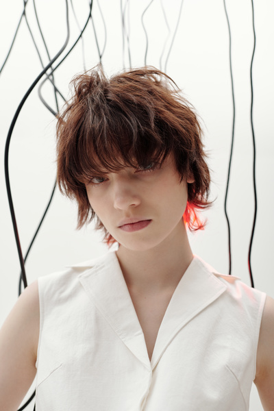 A young woman with short brown tousled hair and strands lighted with red rays dressed in a white sleeveless blouse with a collar stands with her head slightly tilted to the side with black cables hanging behind her on a light background