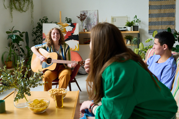 Zoomer friends are sitting on the couch next to the coffee table on which there are snacks and listening to a smiling girl with curly hair sitting on a red armchair playing an acoustic guitar