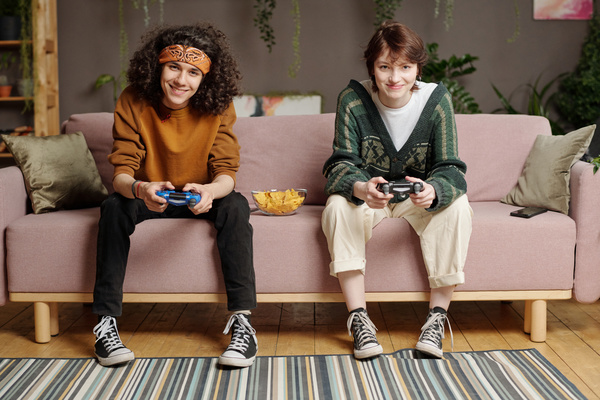 A curly-haired guy in a bandana and a girl with dark short hair in sneakers are smiling sitting on a pink sofa with pillows and a plate of snacks and playing a video game with joysticks