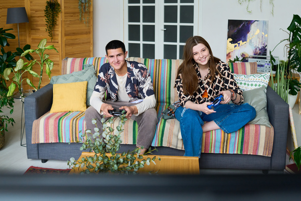 A young guy with black hair in a shirt with a Hawaiian print over a white longsleeve and a girl with long brown hair in a blouse with a pattern are sitting on a sofa with pillows holding joysticks and laughing playing a video game console