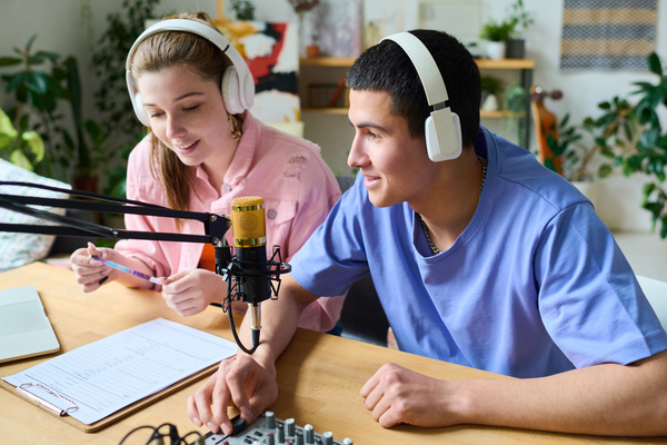 A girl with her hair in a ponytail wearing white headphones and a pink denim jacket holding a pencil in her hands and smiling records a podcast with a young man with dark hair and headphones sitting next to her