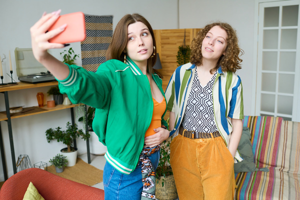 A girl with loose brown hair in a green bomber jacket over an orange top stands next to a curly blonde friend in a striped shirt over a printed T-shirt and takes a selfie on a coral-colored phone