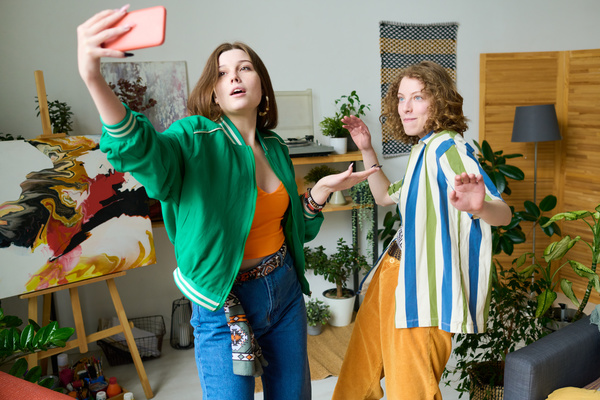 A young woman with brown hair in a green bomber jacket over an orange top standing with a posing friend with short wavy hair in a striped shirt over a printed T-shirt and takes a photo on her phone