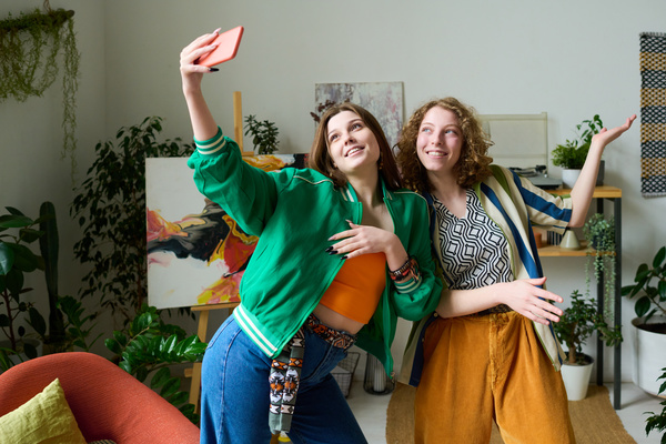 Two young girls in fashionable bright clothes are smiling in a room with plants and a picture and taking selfies posing