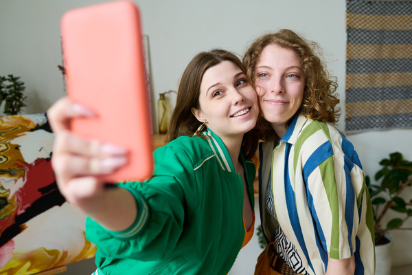 A girl with brown hair in a green bomber jacket takes a photo on her phone leaning cheek to cheek with her friend with curly blond hair and a striped shirt with short sleeves