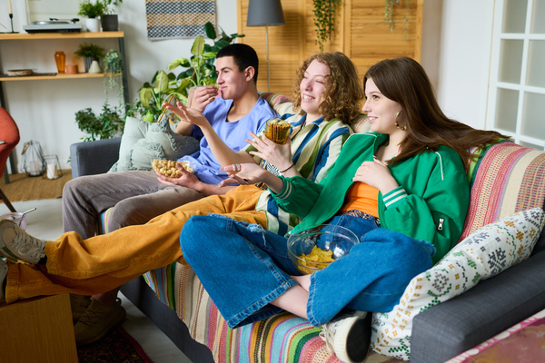 Two young girls in bright outfits are eating bread straws and Sid's chips in relaxed poses on a sofa with a blanket and pillows with their friend who is eating popcorn and watching a TV series with them