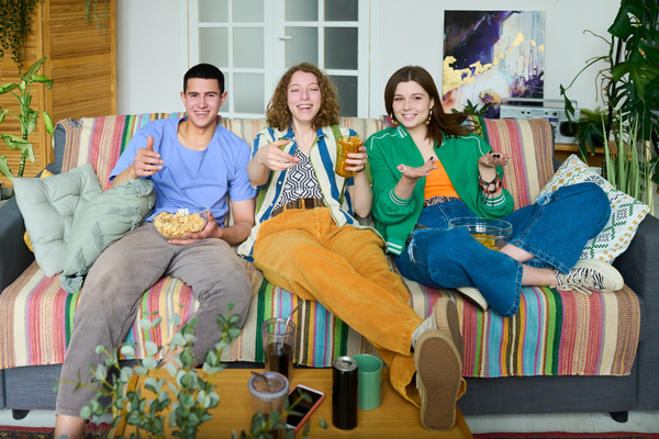 A group of friends in bright youth outfits are sitting relaxed on a sofa with a multicolored striped plaid and pillows in a bright room with plants and laughing watching TV and eating snacks