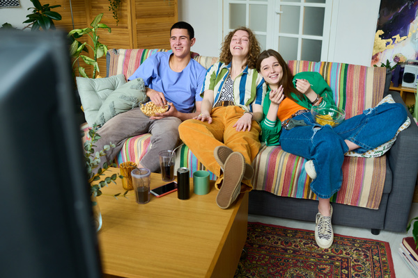 A group of zommers dressed brightly are sitting in the living room on a gray sofa with a striped plaid eating popcorn and chips and laughing at the TV series they are watching