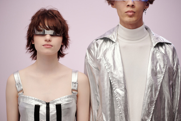 A futuristic couple dressed in fashionable silver-colored outfits and accessories stands straight against of a light pink background