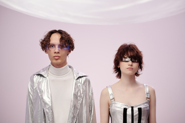 A futuristic man with curly hair in a silver-colored raincoat and a white turtleneck and a woman in a metallic effect dress and glasses stand straight against a light background