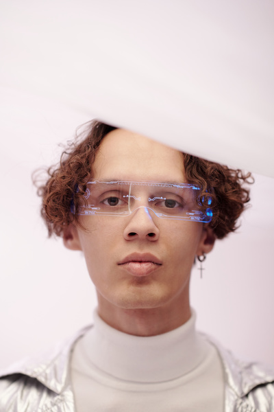 A young futuristic man with curly brown hair in light-colored clothes and transparent glasses with a cyberpunk pattern looks straight ahead