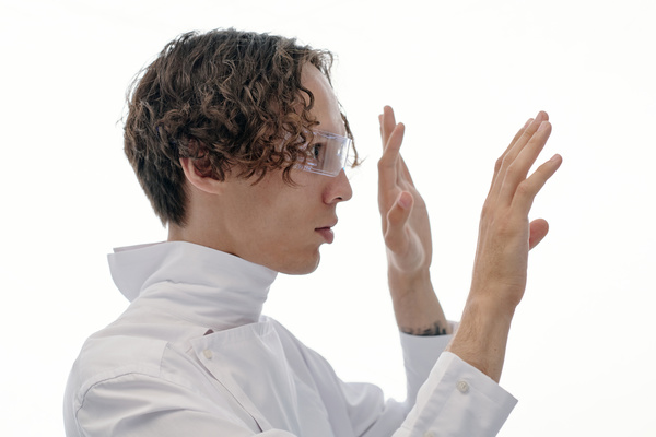 Close-up profile of a man with wavy hair in transparent modern glasses and white clothes with a raised collar looking forward with his hands raised to his face