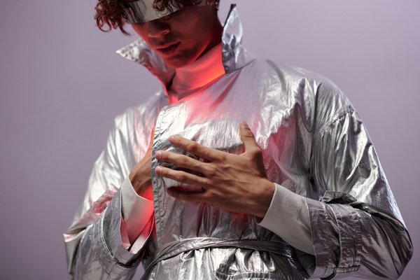 A male cyberpunk lowering his head touches the red glowing heart with his hand which is under his outer clothing