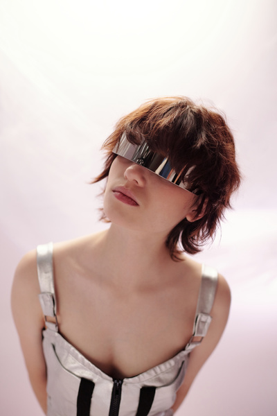 A young futuristic female with short auburn hair dressed in a light dress with straps black elements and a lock on the chest and wearing glasses with one lens hiding her eyes looks up slightly tilting her head to the side