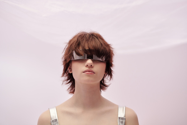 Close up of a futuristic style female in silvery single-lens glasses with short brown hair on a light-pink background