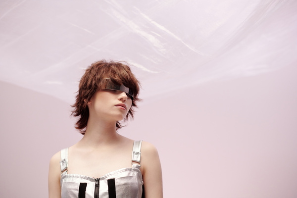 A young woman in a modern silver-colored dress with black stripes and a chest lock in metal single-lens glasses with short brown hair on a light-pink background stands with his head turned to the right