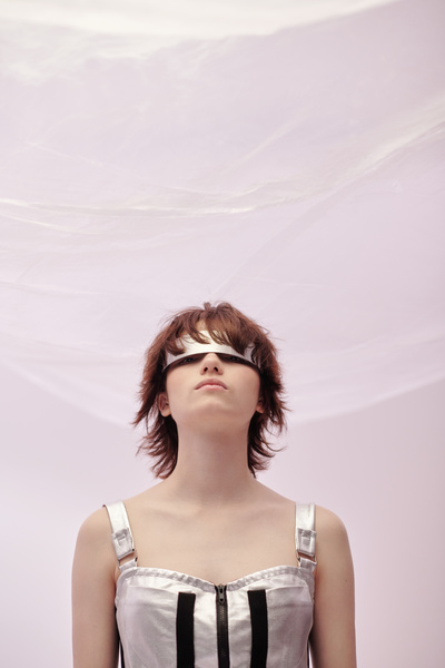 A young futuristic woman in a modern silvery dress with black stripes and a lock on her chest wearing metal glasses with one lens with short brown hair raising her head looks out from under the glasses on a light pink background