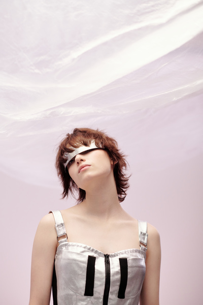 A young female in a modern silvery colored dress with black stripes and a chest lock in metal single-lens glasses with short brown hair on a light-pink background looks out from under the glasses with her head turned to the left