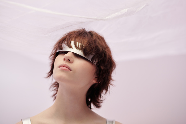 A close-up of a futuristic woman with short wavy brown hair wearing silvery glasses with one lens looking out from under them slightly tilting her head on a light pink background