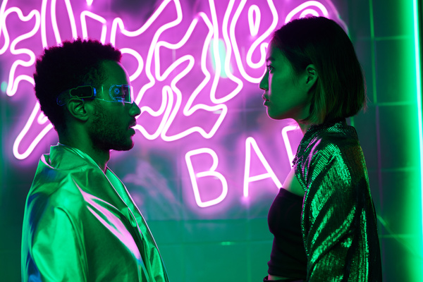 A young man with beard wearing cyber-print glasses dressed in a reflective mantle stands in front of a girl with a short haircut wearing a cape with sequins over a black top against a wall with neon pink drawings in a green-lit bar
