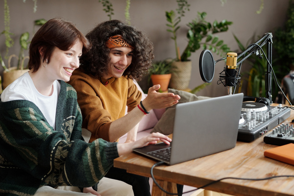 Young people with dark hair in clothes of muted shades laughingly look at the screen of a laptop standing on a table with musical equipment in a room with gray walls and green plants in flower pots