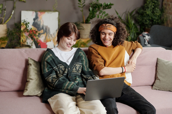 A girl with short dark hair in a green cardigan and light pants is laughing sitting on a pink sofa with pillows with a guy with curly hair in a mustard-colored sweatshirt with a laptop on her lap