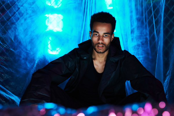 An urbanistically stylized young man with a short Afro hairstyle dressed in a black outfit sits with pink lights in front of him with his hands on his knees against a background of cellophane film illuminated by blue light