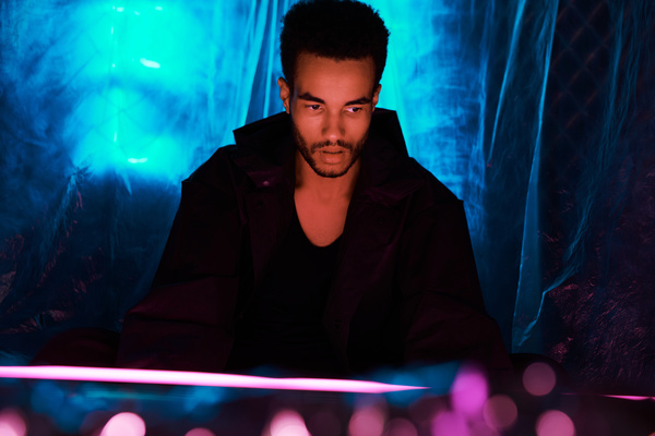 A young man with a short Afro hairstyle dressed in an urban style black outfit is sitting near a pink light source thoughtfully looking at it against a background of cellophane film illuminated by blue light