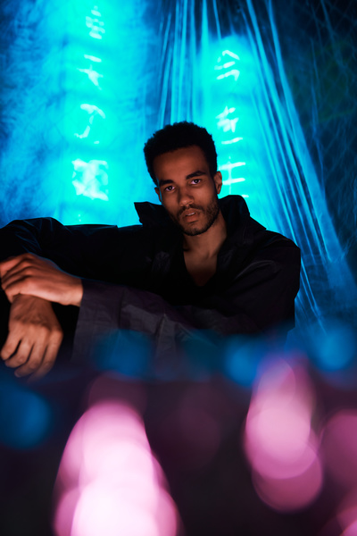A young man with black hair dressed in an urban style black jacket is sitting with his elbow on his knee against a background of blue neon hieroglyphs covered with cellophane film