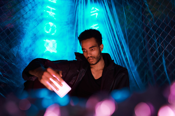 An urban fashioned man with dark curly hair dressed in a black jacket sits with his hand on his knee holding a rectangular source of pink light against a background of blue neon hieroglyphs hidden by cellophane film