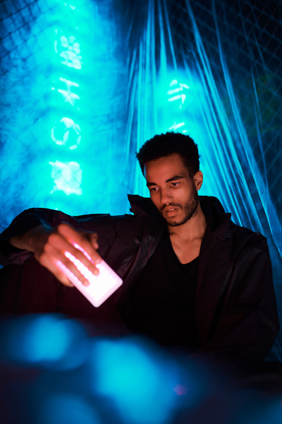 An urban fashioned man with short afro hair dressed in a black jacket over a black T-shirt sits with his hand on his knee holding a pink-light LED lamp against blue neon signs hidden by cellophane
