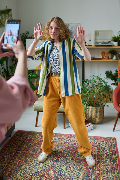 A young curly-haired blonde in a striped shirt and yellow pants poses with her palms out in front of a friend taking pictures of her standing on a Turkish carpet in a room with a lot of greenery in flower pots