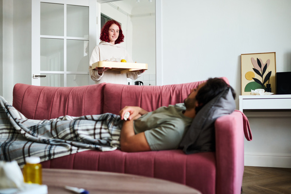 A girl with short dyed red hair dressed in a light hoodie brought breakfast on a tray to a bright living room with a pink sofa on which a sick man with dark hair and beard covered with a blanket is lying