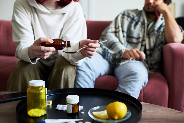 The girl pours medicine for a sore throat into a spoon for her friend who has caught a cold sitting with her who is wrapped in a blanket on the sofa next to which there is a table with a tray of pills and lemon