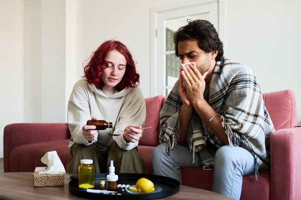 A woman with red hair sitting on the couch with a sick guy who covered his nose with a handkerchief and pours cough syrup taken from a table with medicines and lemon into a spoon