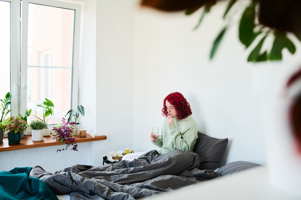 A young girl with a cold with red and wavy hair in a bright room with flowers on the windowsill sitting on the bed and covered with a black blanket takes a pill and is going to drink water