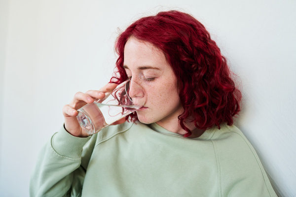 A young sick woman with freckles on her face and bright red hair in a green hoodie against a white wall drinks a pill with a glass of water