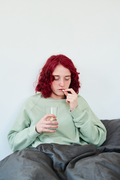 Covered with a black blanket a sick girl with red and wavy hair is sitting behind a white wall about to take a pill and drink water