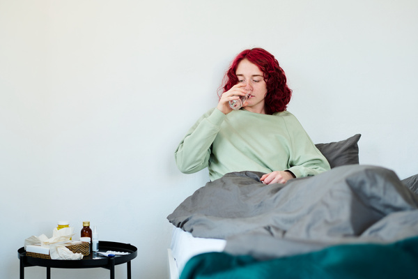 A woman with a cold with short red hair dressed in light pajamas is sitting on the bed next to which there is a table with paper handkerchiefs and medicines covered with a gray blanket and drinking water