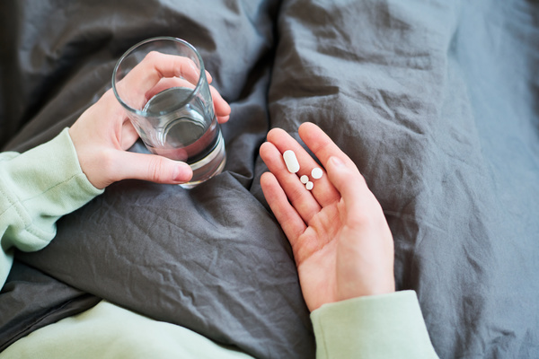 A woman with a cold wrapped in a gray blanket holds a handful of medicines and a glass of water in her palm