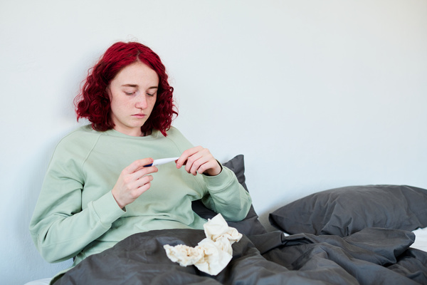 A girl with red hair in green clothes sitting in a dark bed taking her temperature and looking at a thermometer