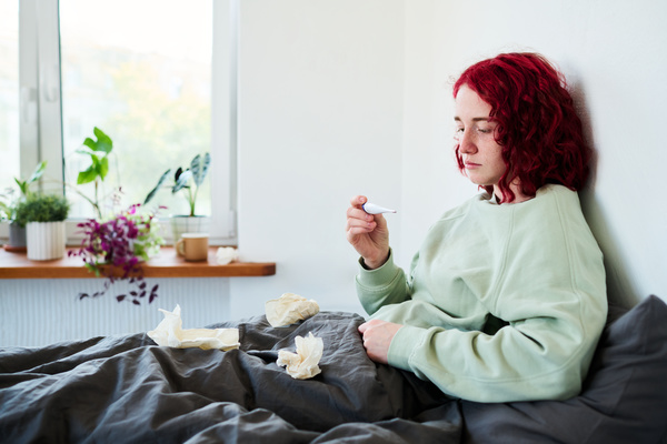 profile of a young woman with freckles sitting on a bed with napkins on it and measuring the temperature against the background of a white room with a window