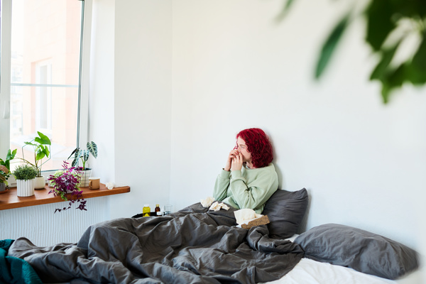 A illgirl with red curly hair in home clothes is sitting in a gray bed with a box full of paper napkins and blowing her nose in a handkerchief in a bright room with flowers on the windowsill