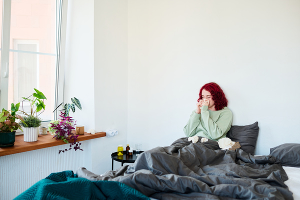 A young woman with bright dyed hair sitting in bed with crumpled handkerchiefs around pressing one of them to her clogged nose in a room with greenery standing on the windowsill and a table full of medicines