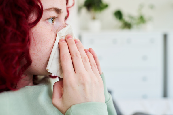 A sick girl with burgundy hair and freckles dressed in light pajamas cleans a clogged nose by pressing a white napkin to it