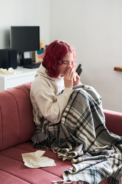 A young woman with red curly hair in a white hoodie caught a cold and sitting on the couch with napkins covered with a plaid blanket cleans her nose inside by pressing a handkerchief to it