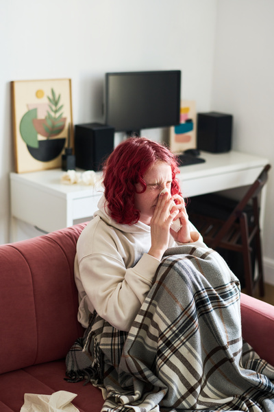 A girl with bright dyed wavy hair in a white hoodie got sick and sitting on the sofa in the living room wrapped herself in a plaid with a print and wincing blows her nose into a handkerchief