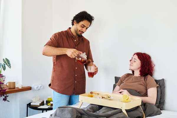 A guy with black hair and beard in a shirt pours tea to a girl with a cold who is sitting in a gray bed with a tray on which lies lemon and ginger