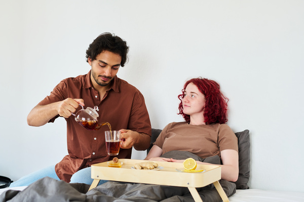 A man in a shirt pours tea to a sick woman sitting with her on the bed next to a wooden tray on which there is lemon ginger and honey