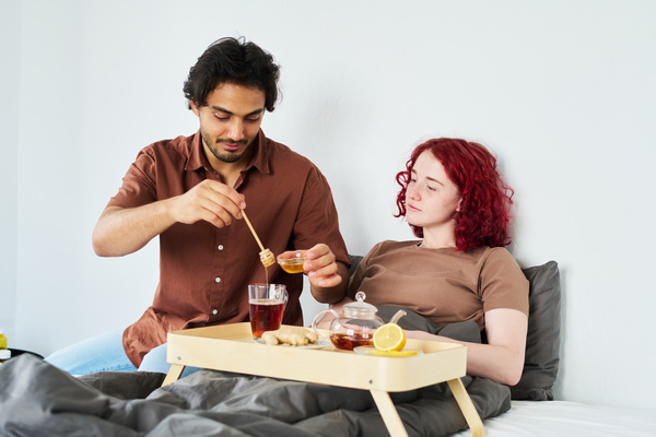 A guy with dark hair and a beard adds honey to tea for his sick girlfriend with brightly dyed hair who is sitting next to him in bed